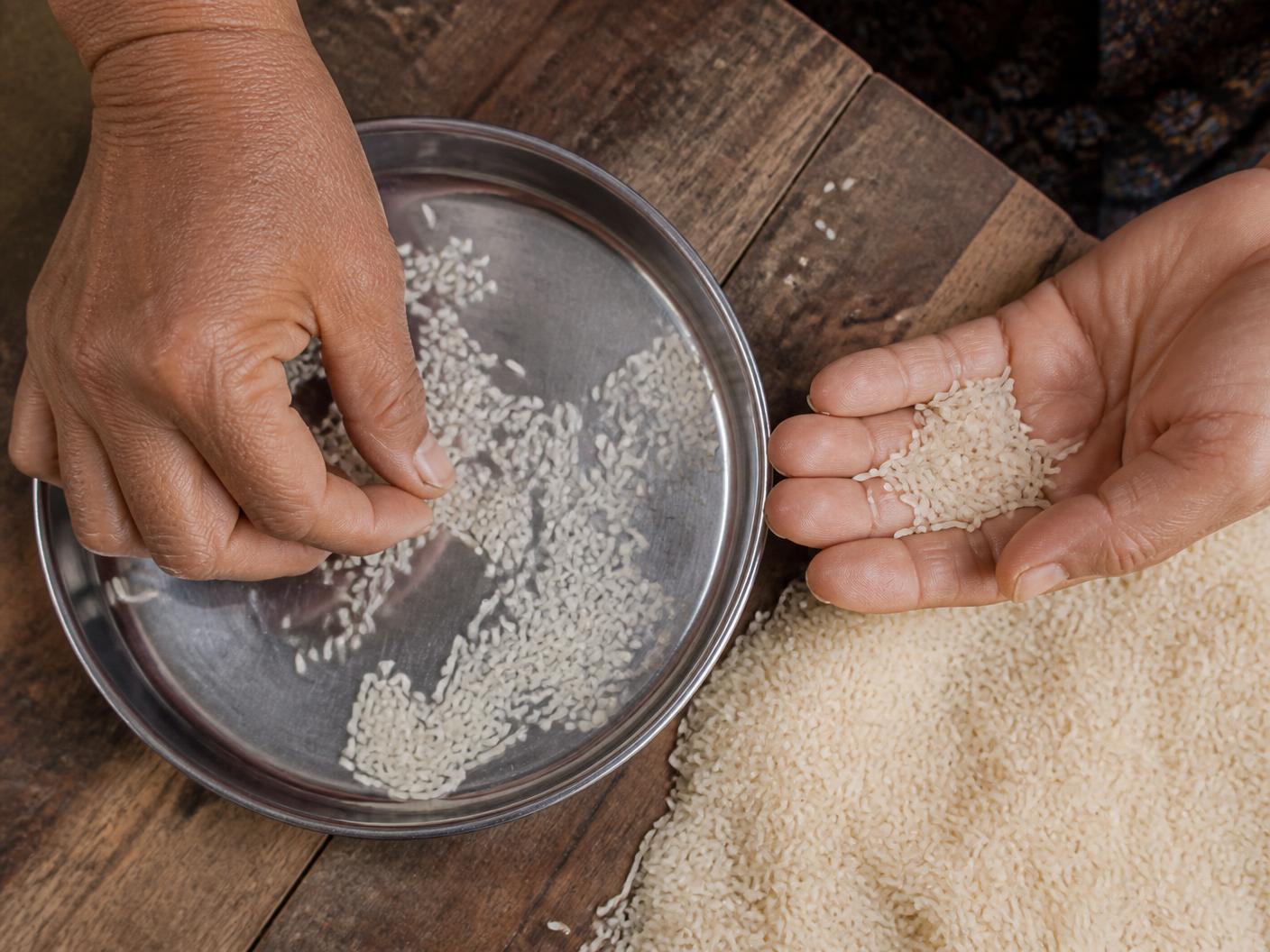 Hands inspecting white rice grains on a tray over a wooden table